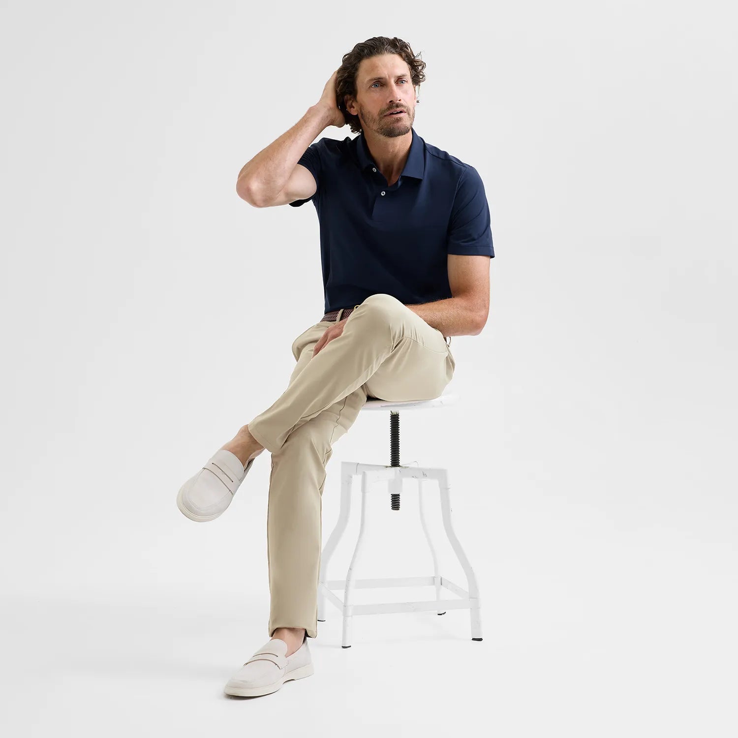 Man sitting on a stool wearing a navy como polo shirt, sand Positano pants, and bone loafers against a white background