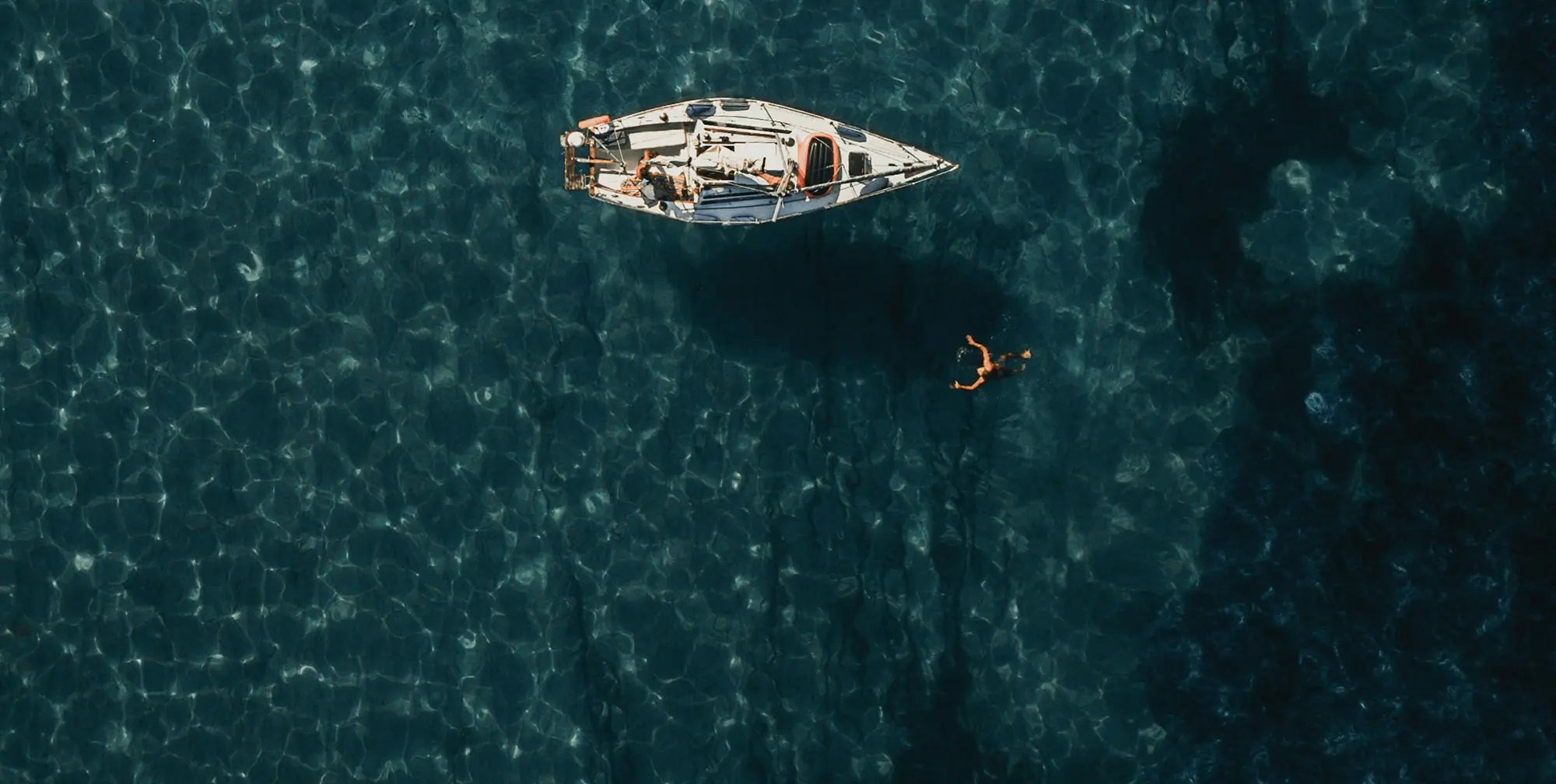 Man swimming next to sailboat – epitomizing freedom and adventure, with stylish, performance-driven menswear for the modern traveller.